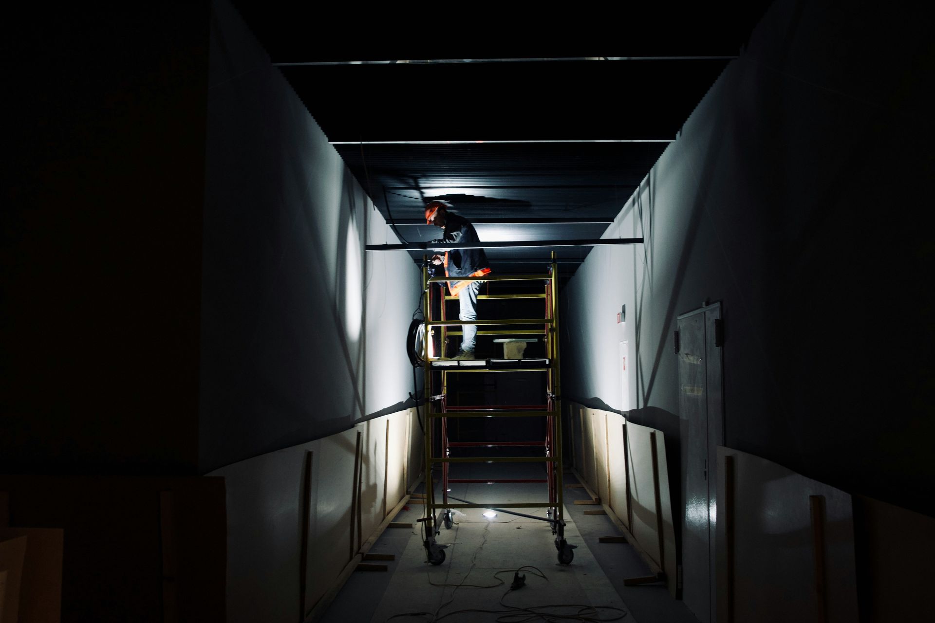 Worker on a scaffold in a dim hallway, installing ceiling panels with a flashlight.