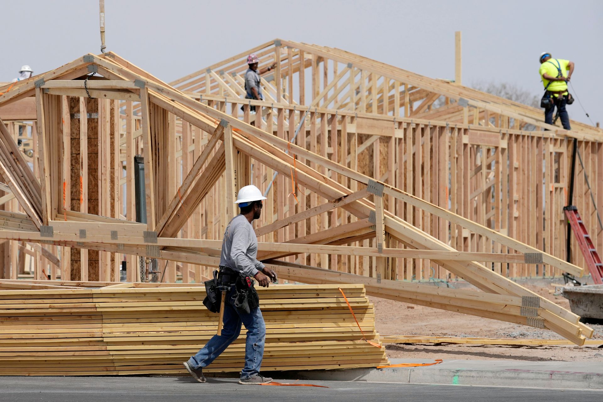 Construction workers framing a house with wooden beams on a site, one carrying lumber in front.