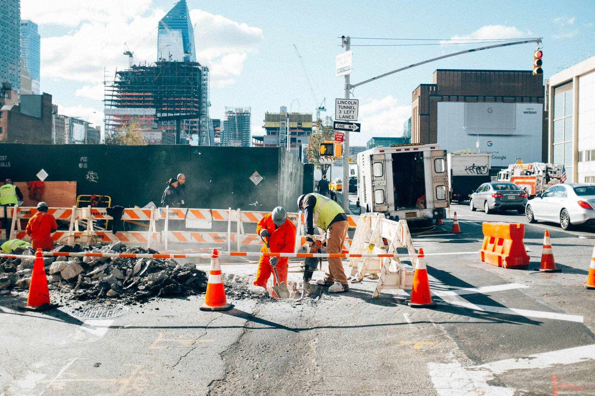 Road construction crew repairing a city street with orange cones, barriers, and traffic in the background