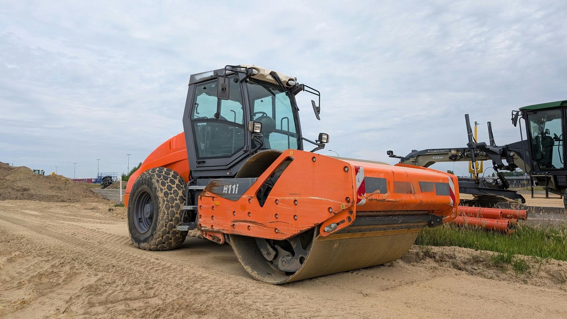 Orange road roller parked on a dirt lot beside another heavy construction machine