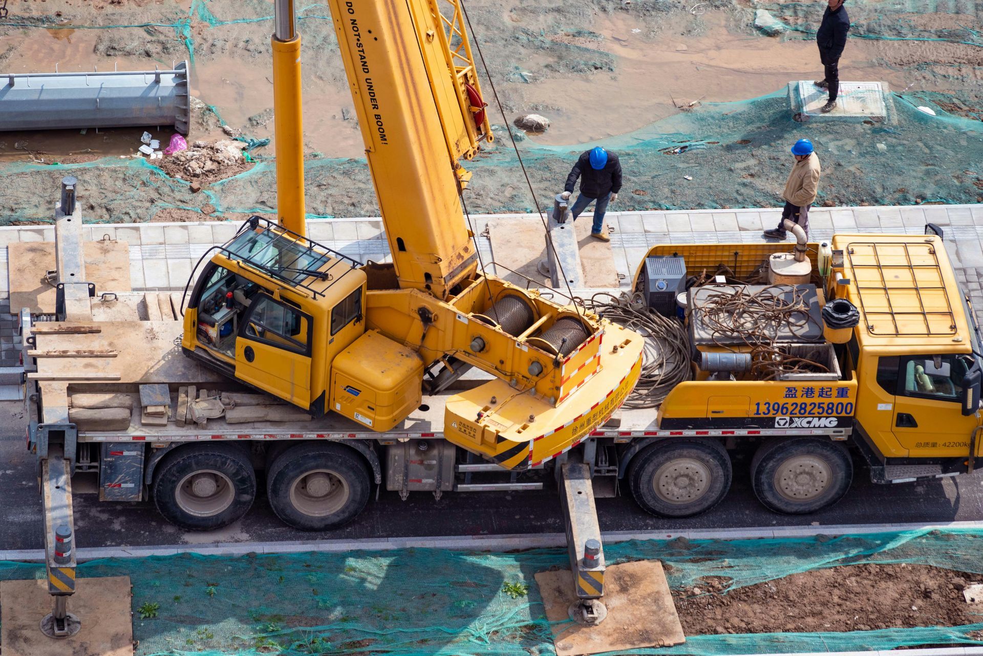 Yellow crane loaded on flatbed truck at a construction site, with workers nearby.