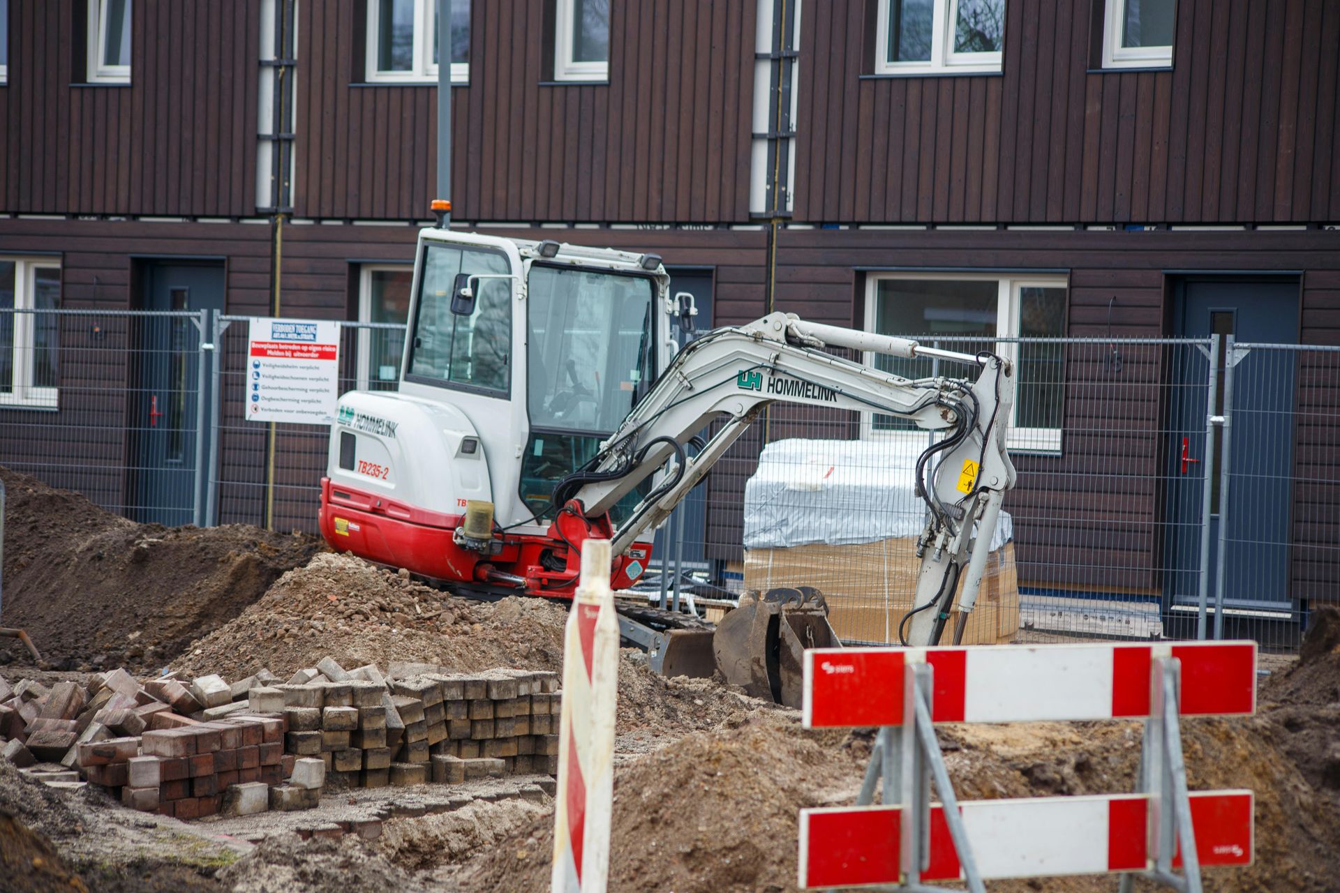 Excavator digging in a construction site beside a brown building, with a red-and-white barrier in front.