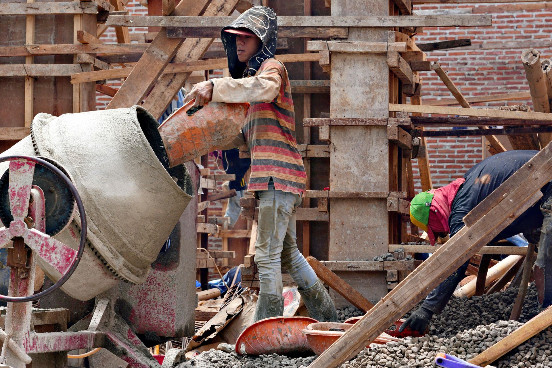 Construction workers pouring concrete at a brick building site, with a cement mixer and wooden scaffolding.