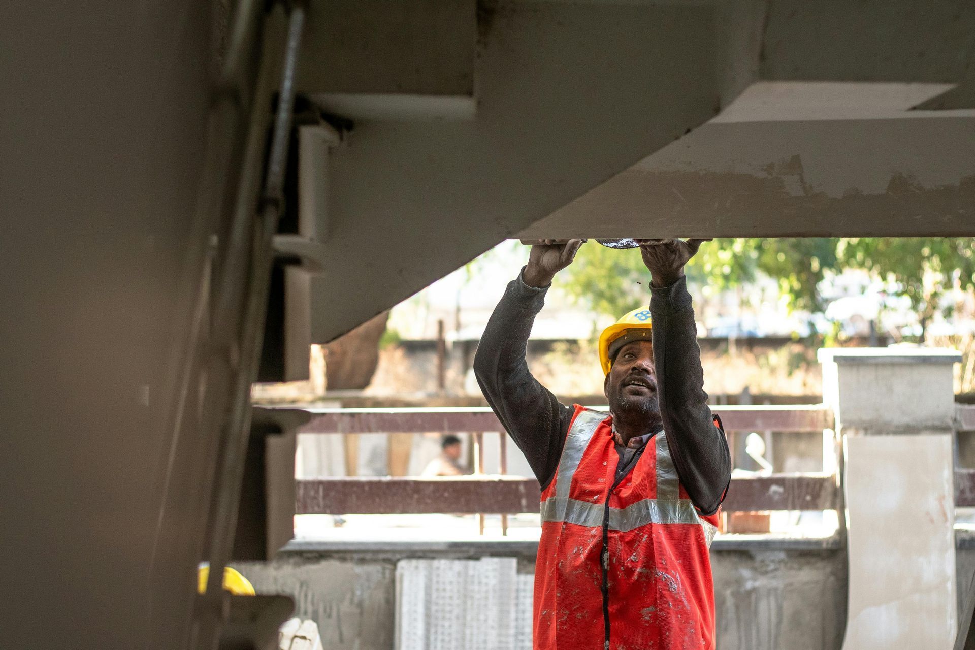 Worker in red overalls and yellow hard hat lifting a beam under a roofed structure.