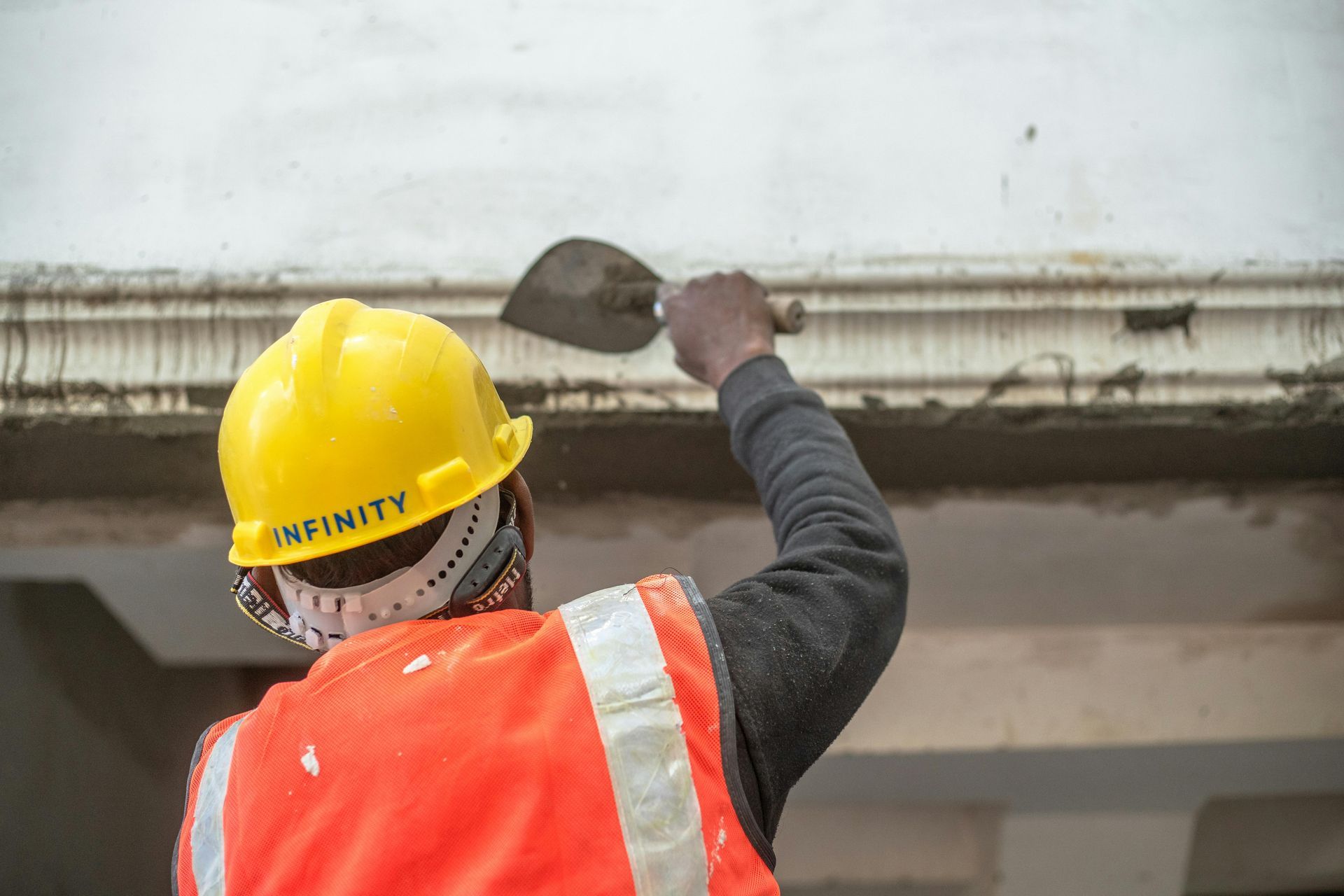 Worker in a yellow hard hat and orange vest scrapes debris from a building ledge with a trowel.