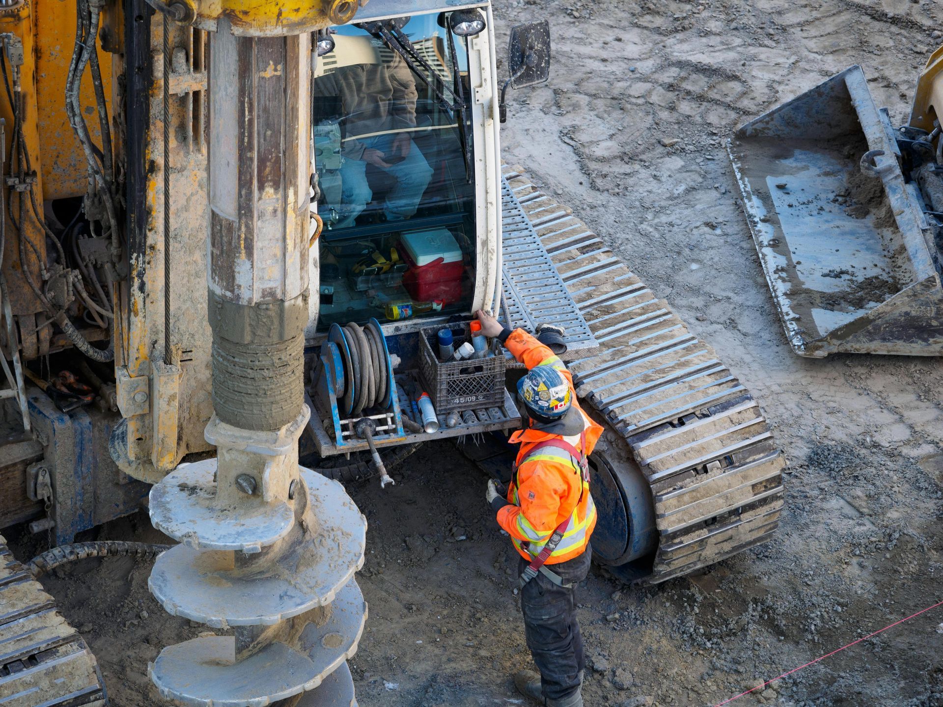 Construction workers inspect a drilling rig beside a large auger on a muddy site.