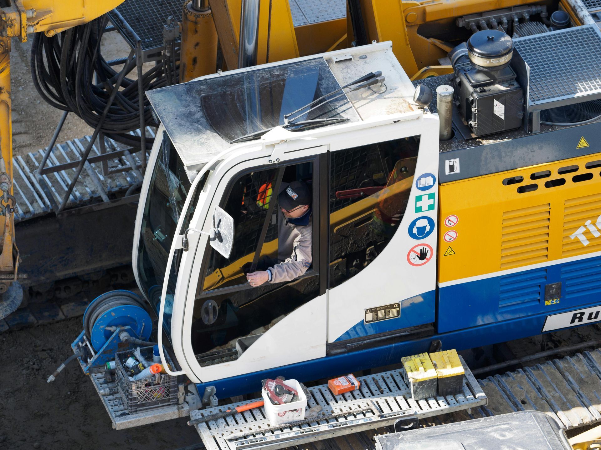 Yellow and white construction machine cab with a seated operator, viewed from above on a worksite.
