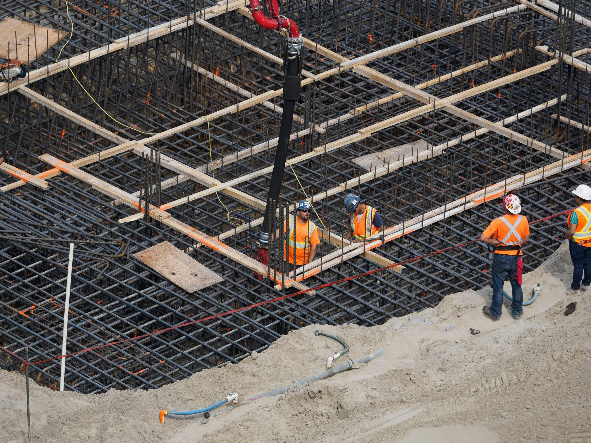 Construction workers in orange vests on rebar foundation with concrete pump hose