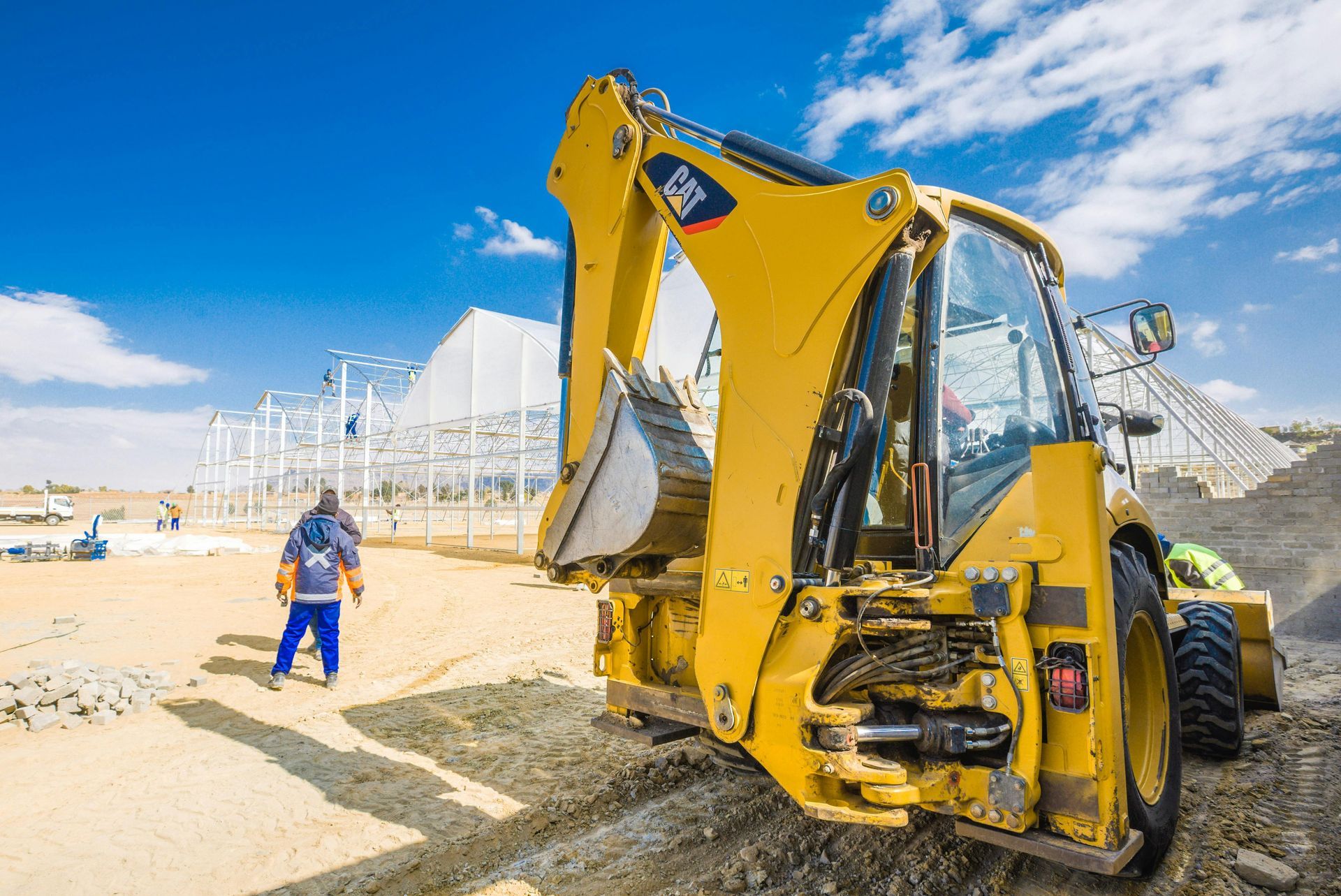 Yellow backhoe loader on a sandy construction site with a worker in blue nearby under a clear sky