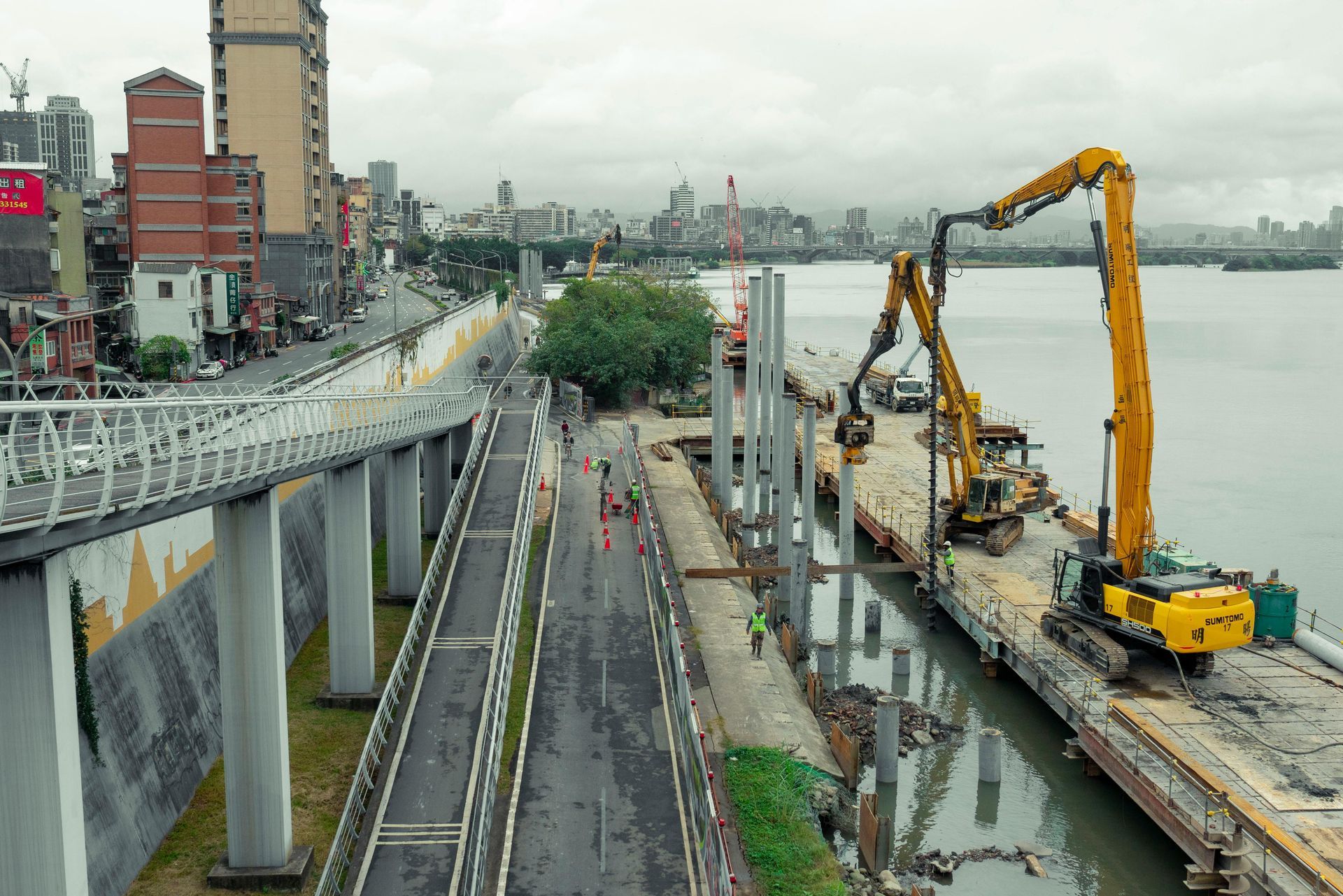 Construction site with excavators beside a riverside road and elevated bridge in a gray cityscape
