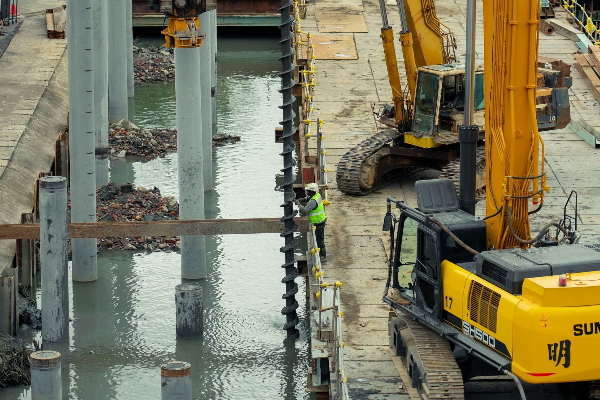 Construction site beside a waterway, with cranes, workers in safety gear, and concrete pillars along the edge