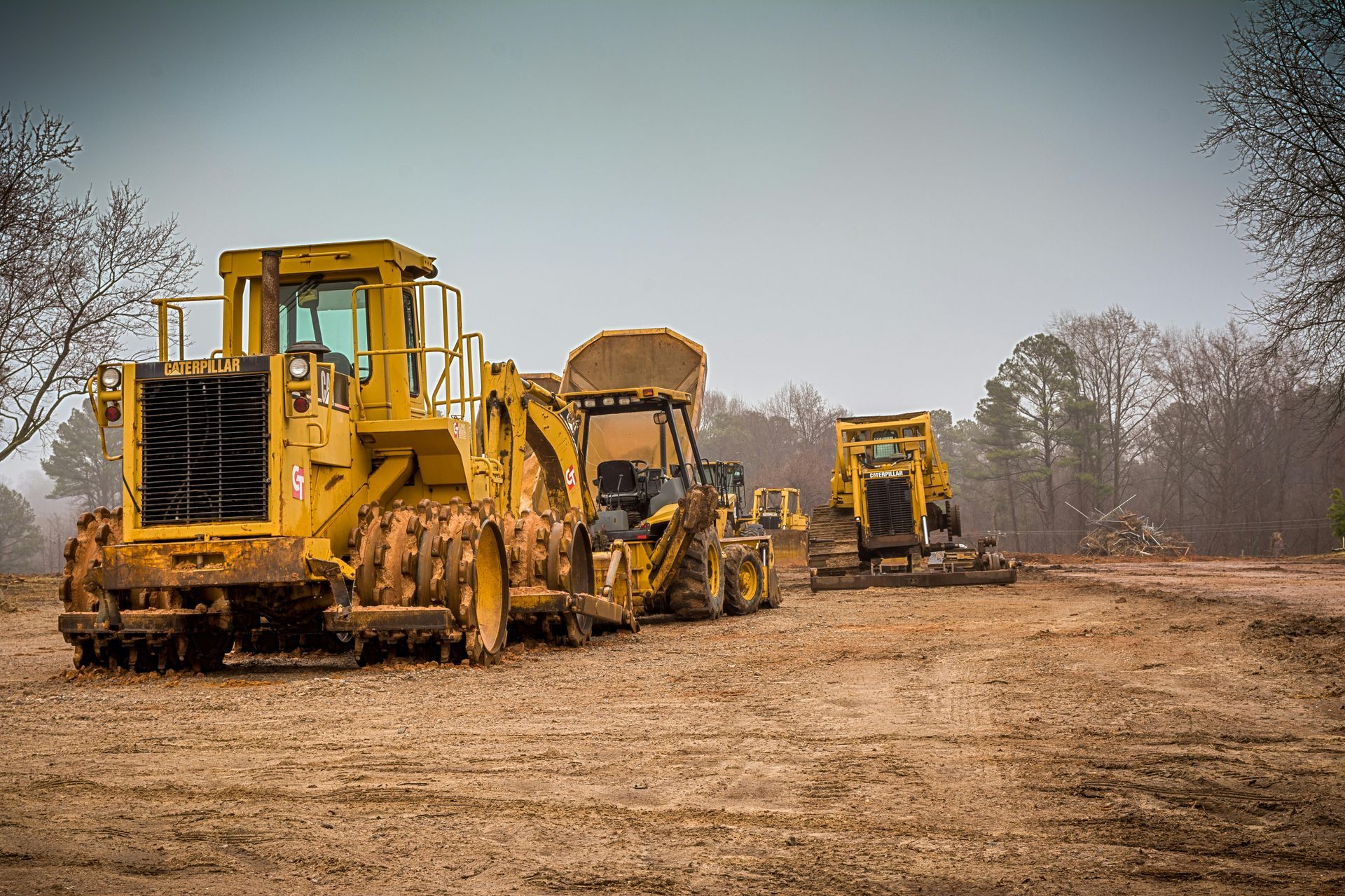 Three yellow construction vehicles on a dirt road beside a wooded area under a cloudy sky