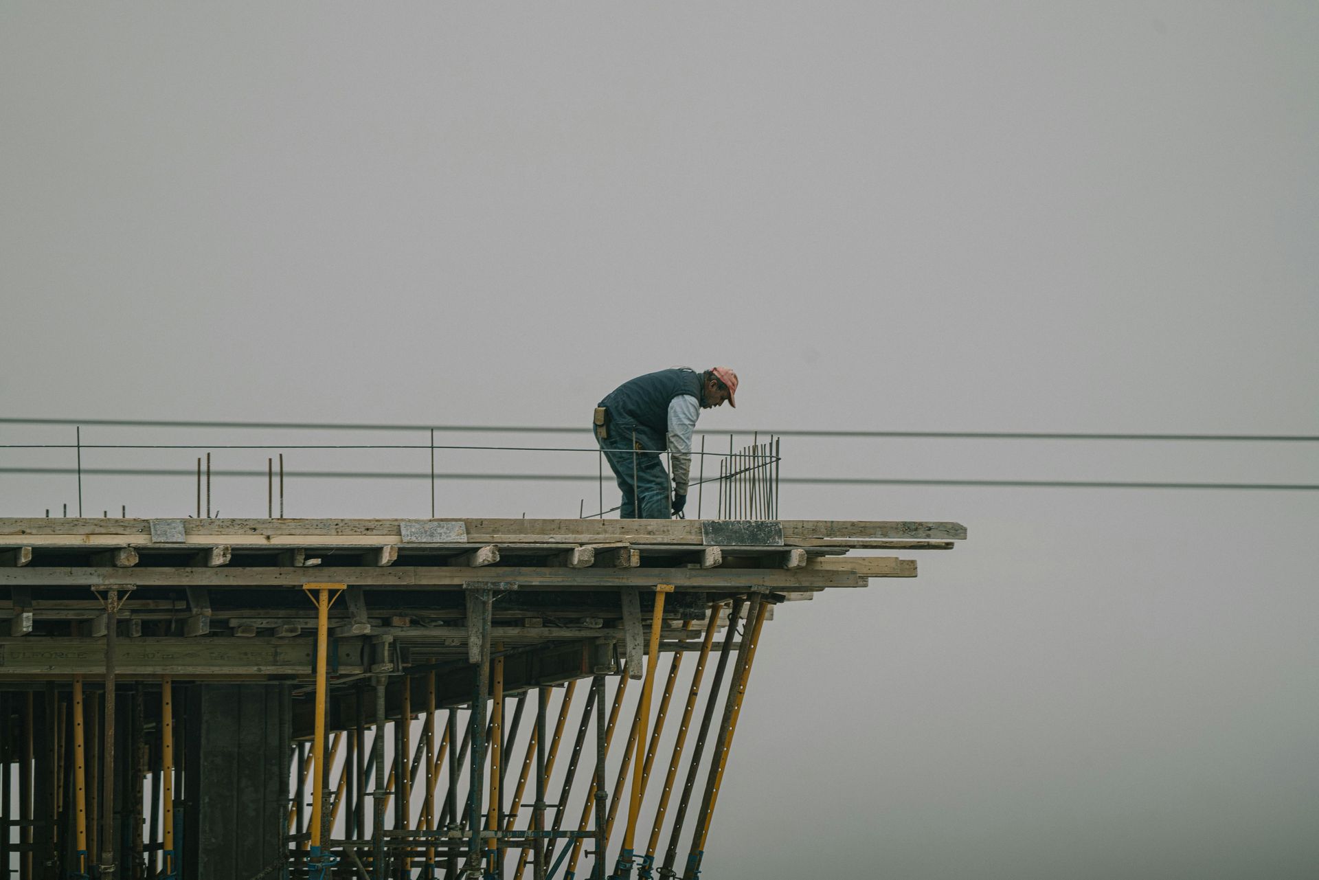 Worker bent over on a construction platform high above the ground, with scaffolding against a gray sky