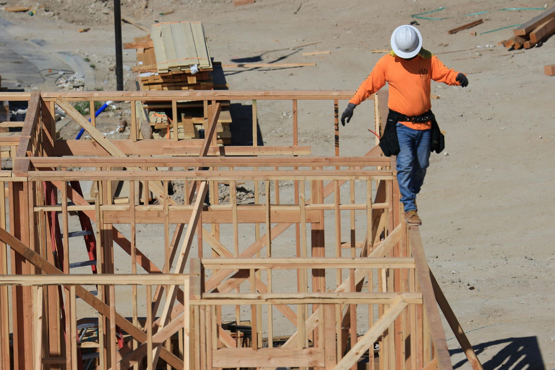 Construction worker in orange shirt and white hard hat walking beside wooden framing on a building site