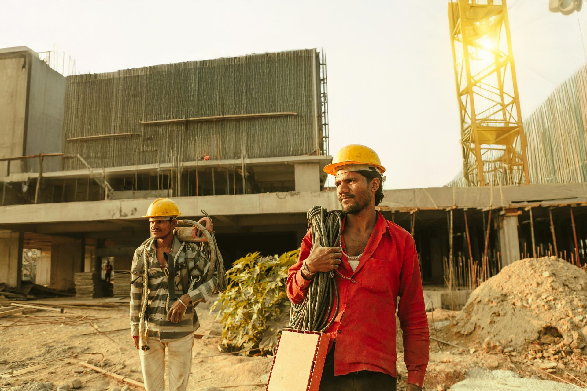 Two construction workers in hard hats walk on a building site beside scaffolding and a crane.