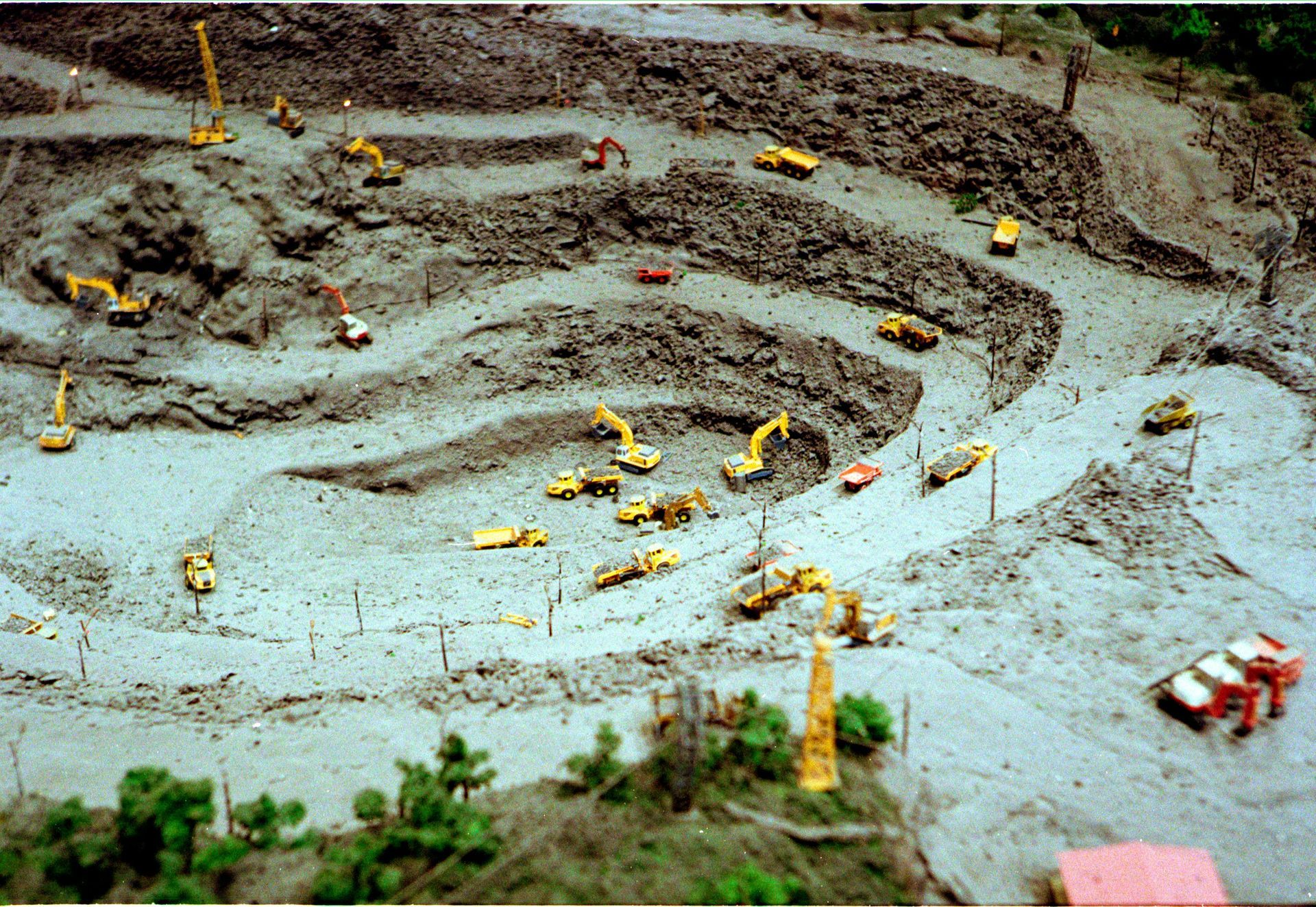 Tiny excavators work in a muddy construction site with terraced earth and scattered vehicles.
