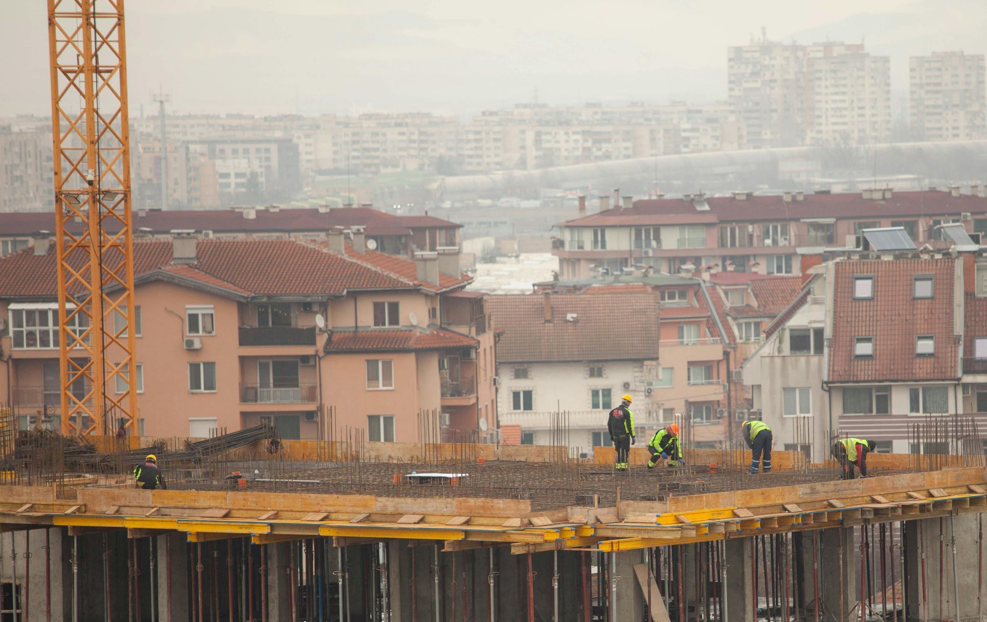 Construction workers on a high-rise slab with a crane, overlooking residential buildings in hazy daylight
