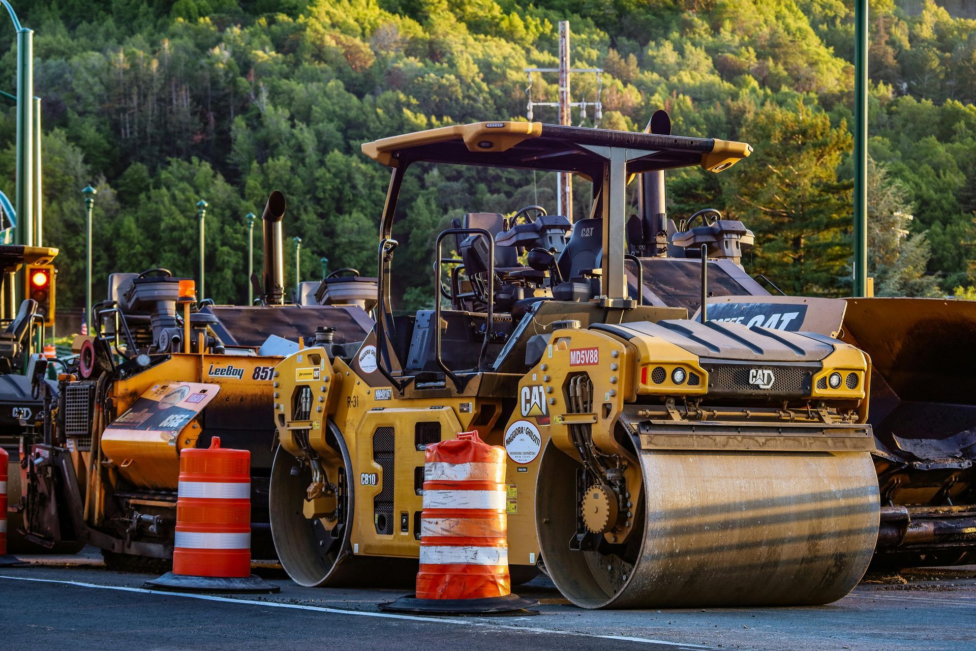 Yellow road rollers lined up behind orange traffic barrels on a roadwork site with wooded hills in the background
