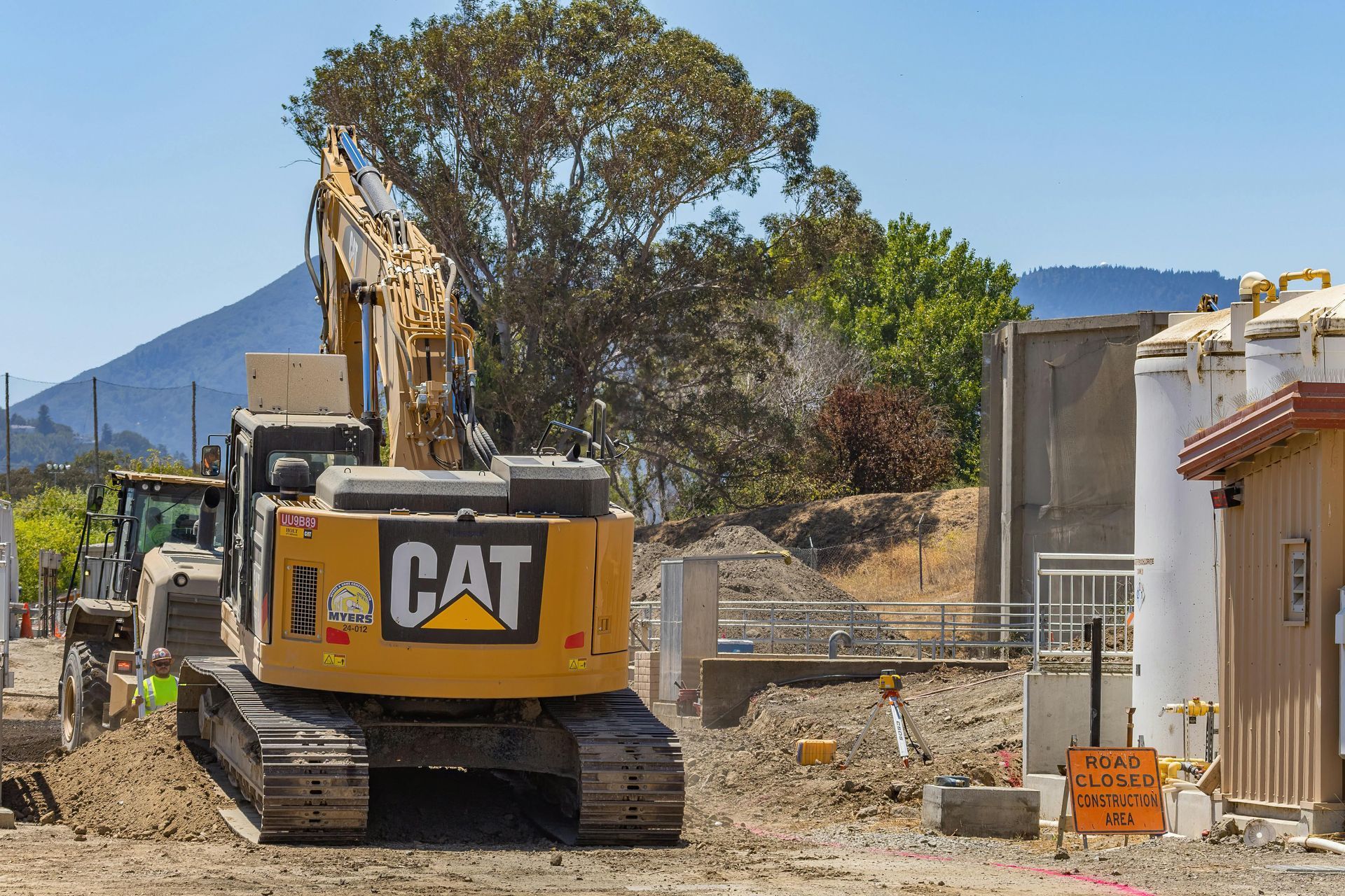 CAT excavator at a construction site beside a house, with dirt piles and trees in the background