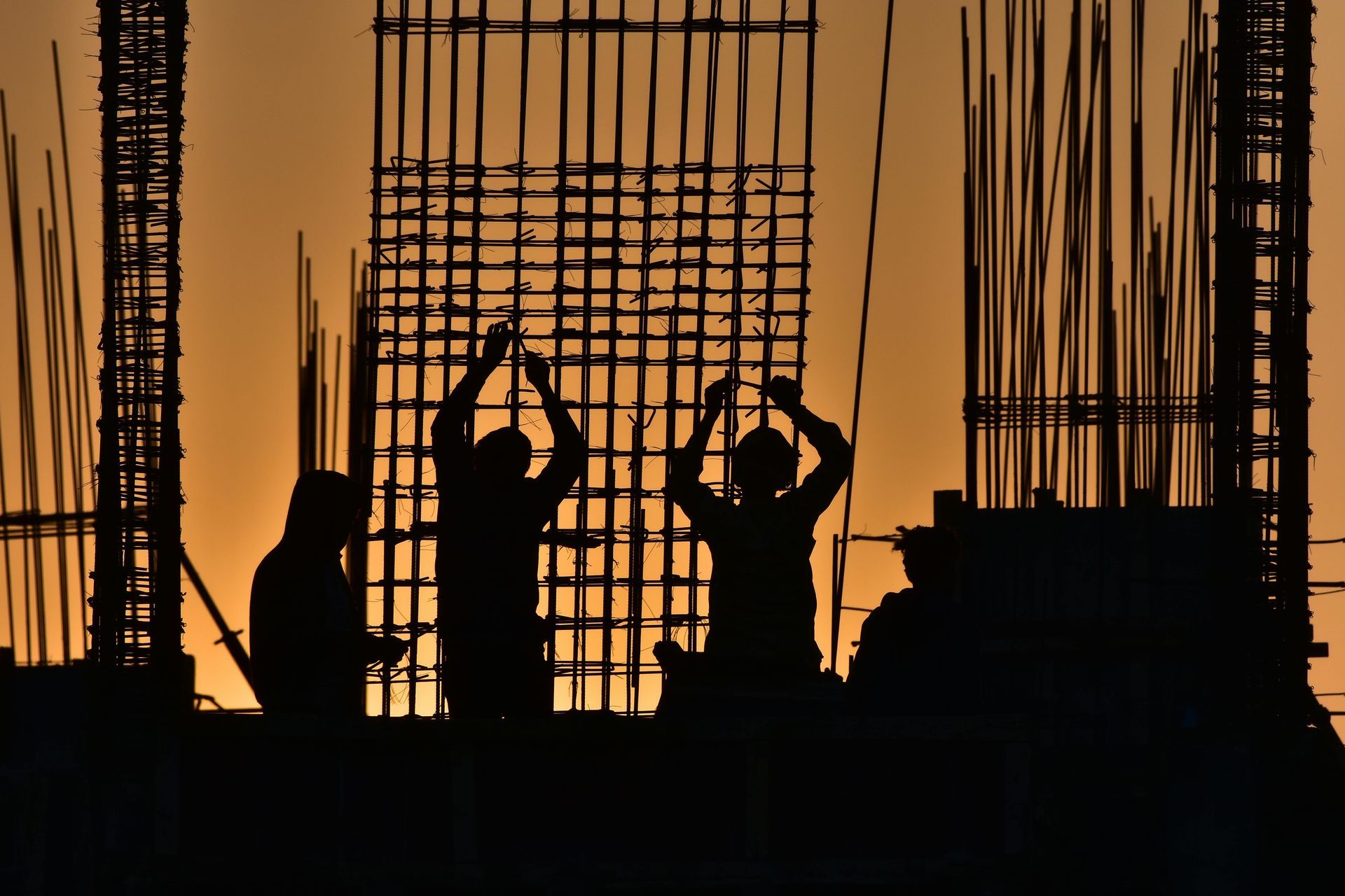 Silhouetted construction workers on a building site at sunset, framed by vertical rebar columns.