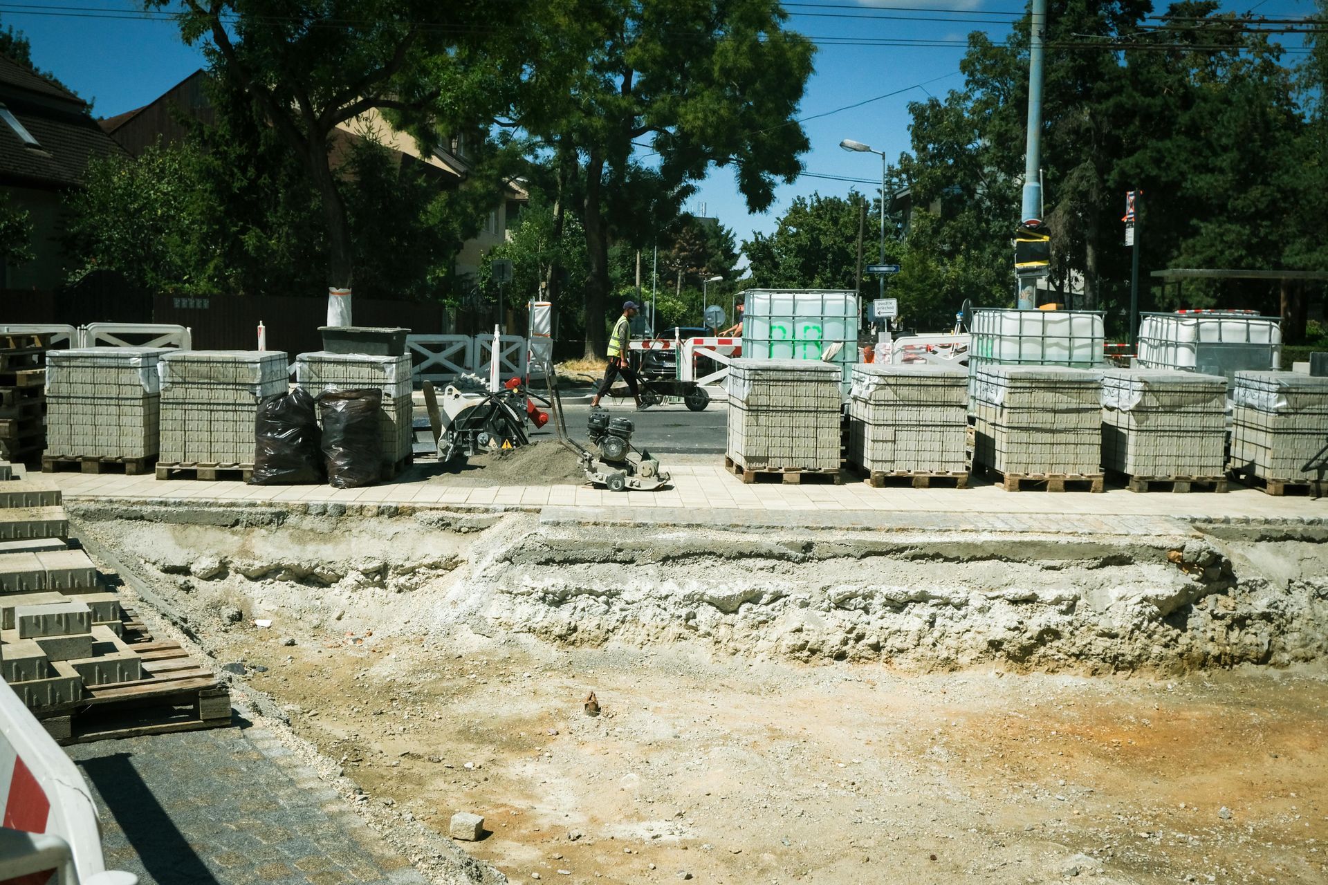 Construction site with stacked concrete blocks, heavy equipment, and a dirt road under trees