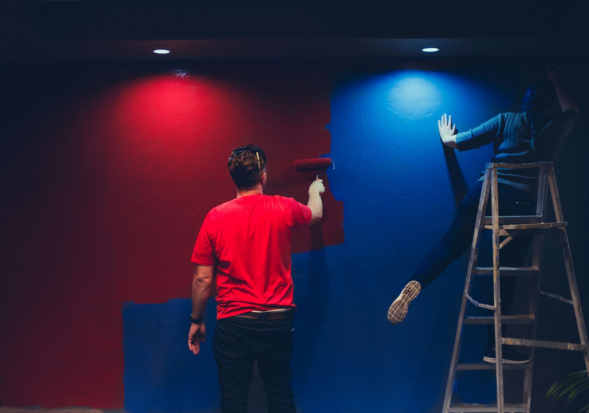 Person in red painting a wall under red and blue lights; another person stands on a ladder.