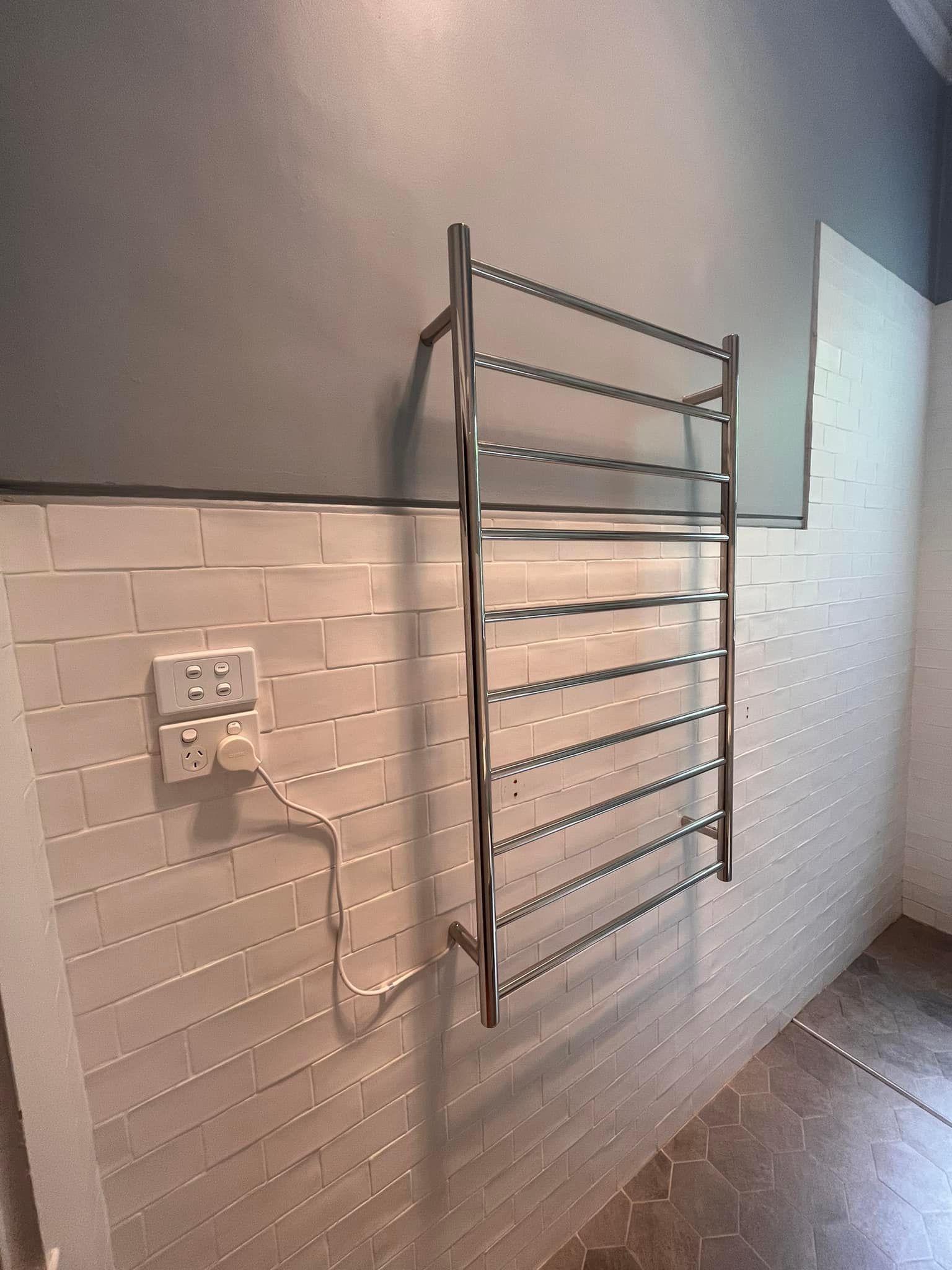 Heated towel rack on tiled wall next to a power outlet.