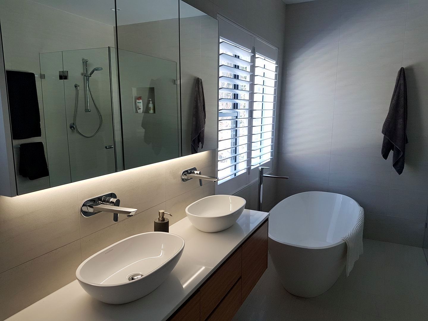 Modern bathroom with dual sinks, a bathtub, and shuttered window.