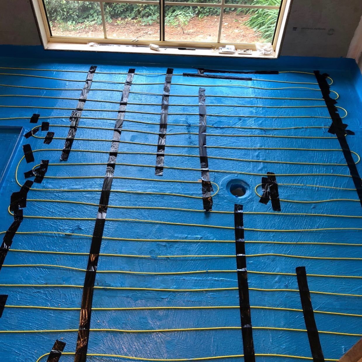 Blue bathroom floor with installed radiant heating cables taped down, near a window and drain.