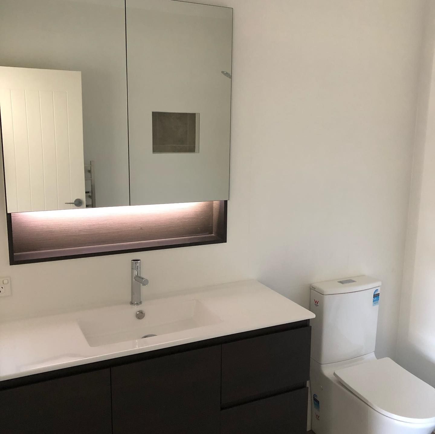 Bathroom with white walls, dark vanity, rectangular mirror with light, and a white toilet.