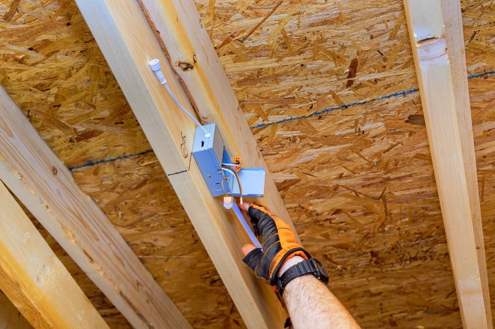 Person installing electrical wiring in a wooden ceiling with a metal box, wearing work gloves.
