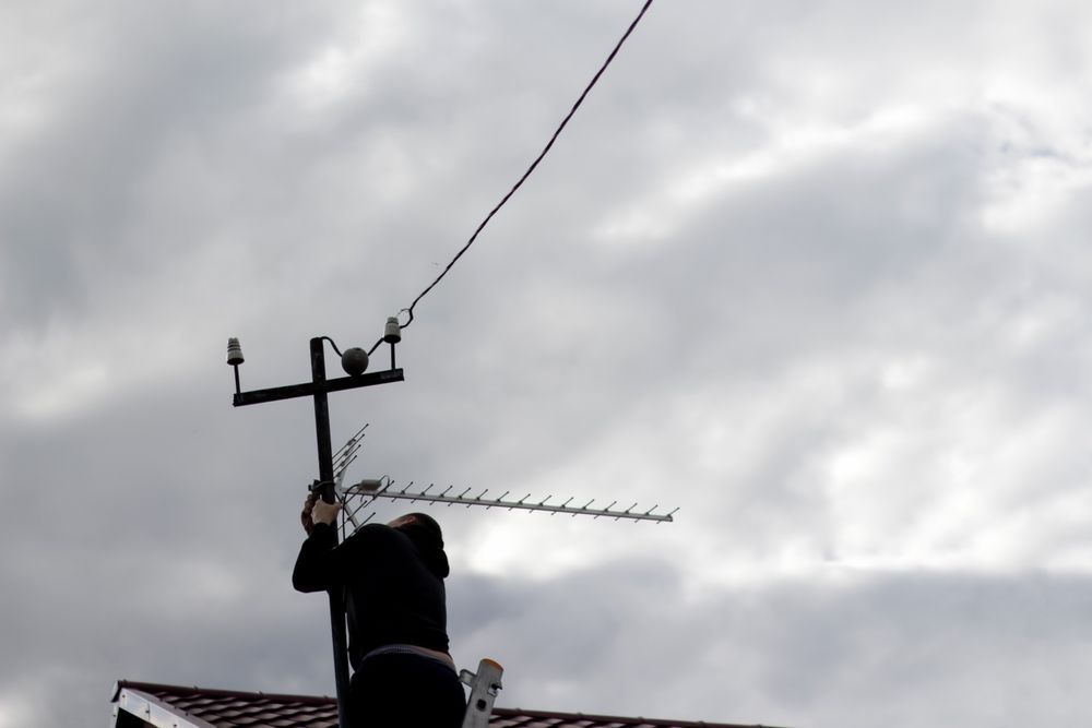 Person on rooftop adjusting an antenna, near utility pole with electrical lines, cloudy sky.
