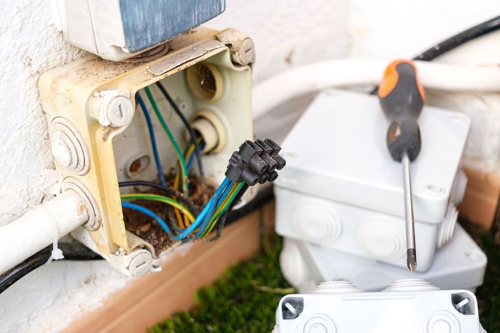 Open electrical junction box with exposed wires, a screwdriver, and additional boxes on a white exterior wall.