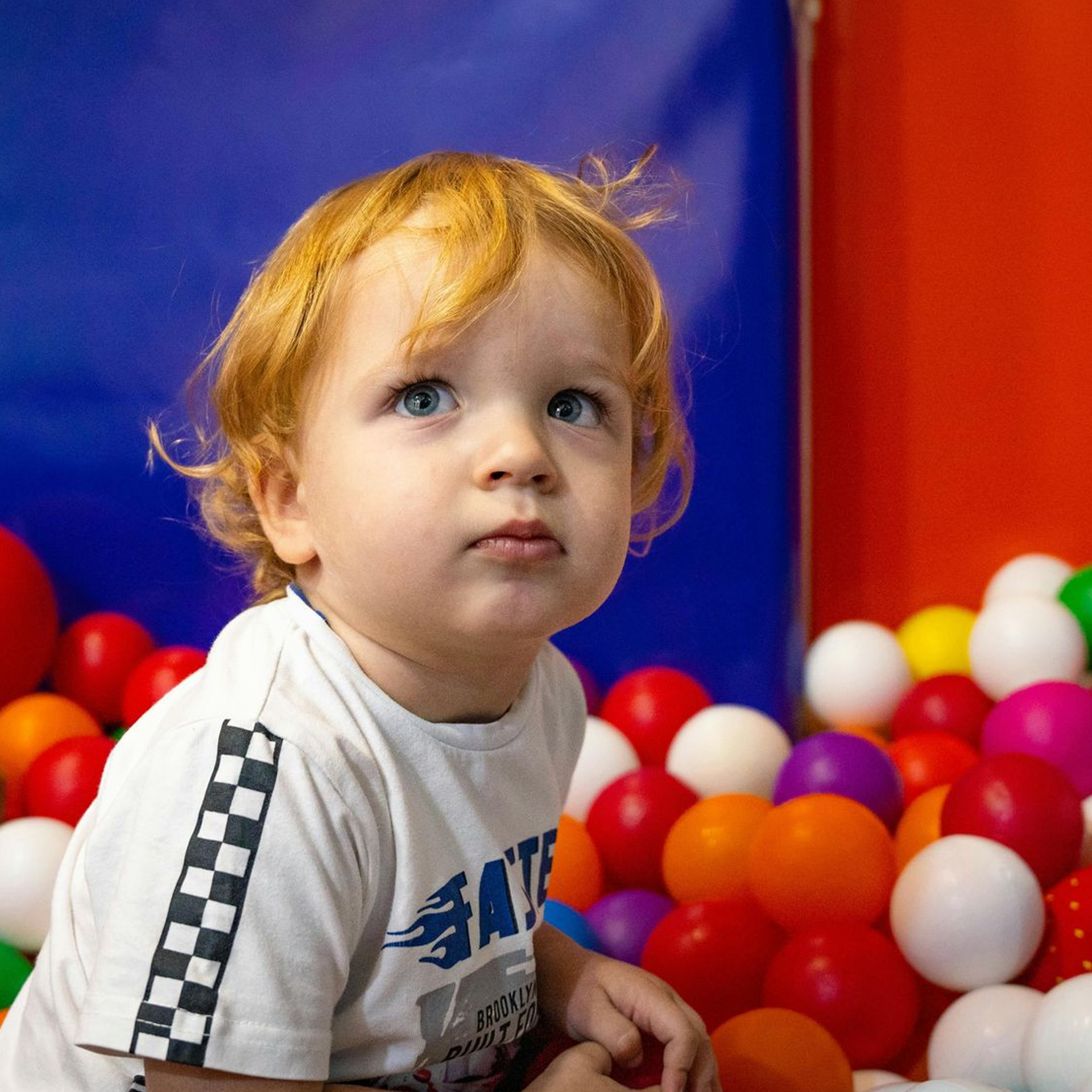Young child with red hair in a ball pit, looking up with a thoughtful expression.
