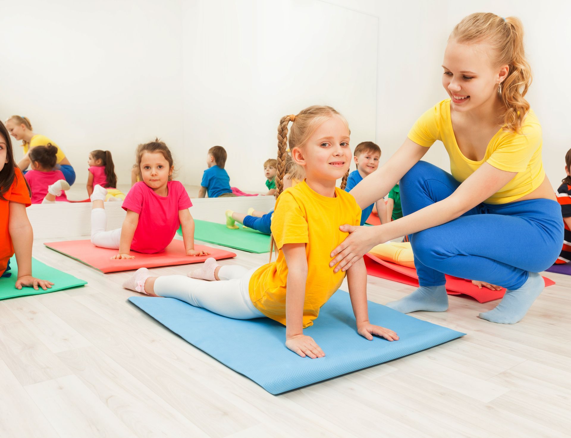 Children and instructor in a yoga class. A young instructor guides a girl in a yoga pose on a blue mat in a bright studio.