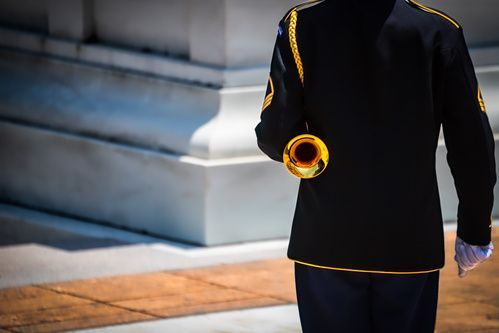 A person in a black uniform with gold trim holds a brass trumpet outdoors near stone steps.