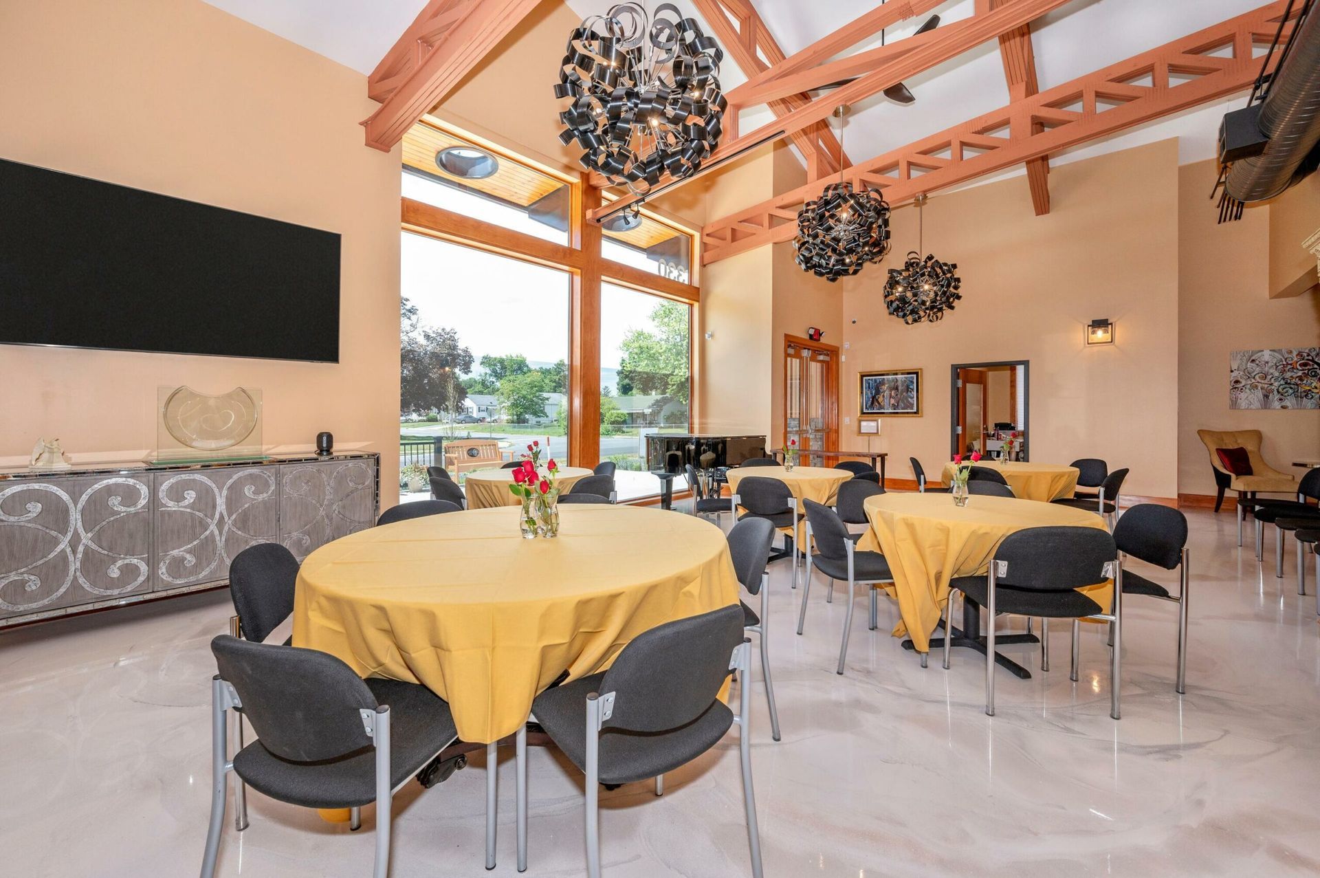 Bright café with yellow tablecloths, black chairs, high ceiling, and large windows overlooking greenery.