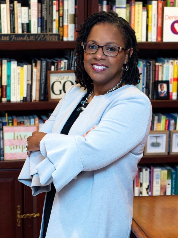 Woman in a light blue blazer standing in front of bookshelves, smiling with arms crossed.