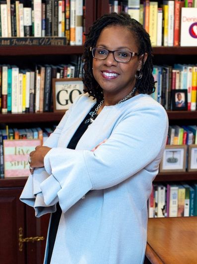Woman in a light blue blazer stands smiling in front of a bookshelf in a library.