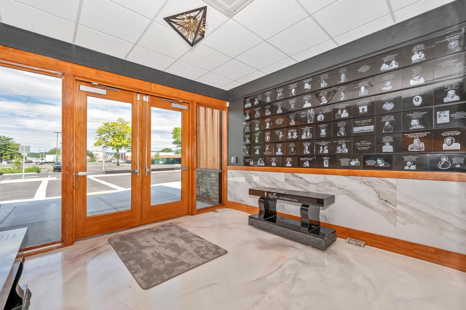 Bright lobby with wood-framed glass doors, dark wall board, and a bench on glossy tile floor