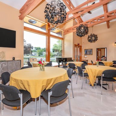 Bright dining hall with round tables covered in yellow cloths, dark chairs, and large windows overlooking greenery