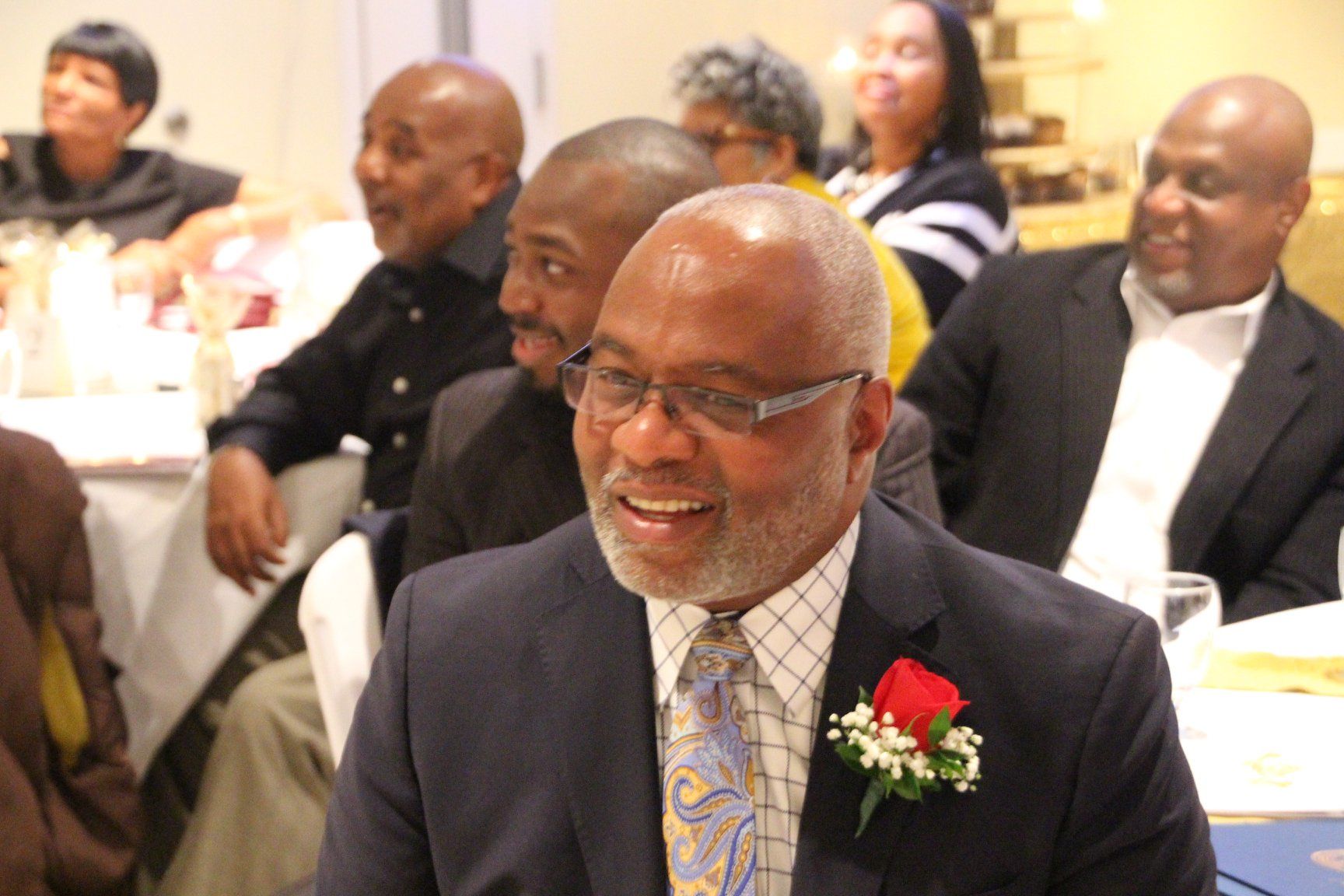 Man in suit with glasses and red boutonniere smiling at a formal banquet table
