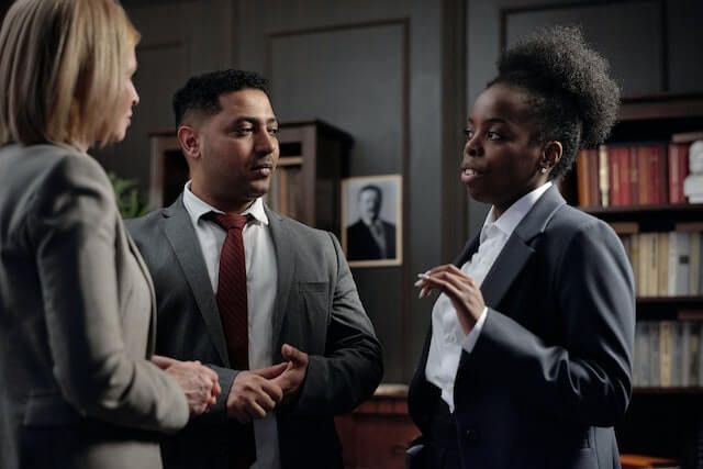 Three people in business attire talking in an office, with shelves and a framed portrait in the background.