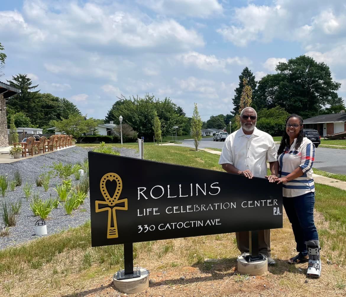 Two people standing beside a Rollins Life Celebrations Center sign in a landscaped garden