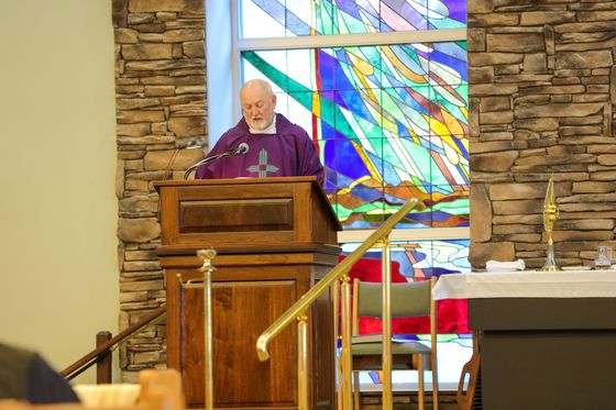 A person in purple liturgical robes speaks from a wooden lectern in front of a colorful stained-glass window.