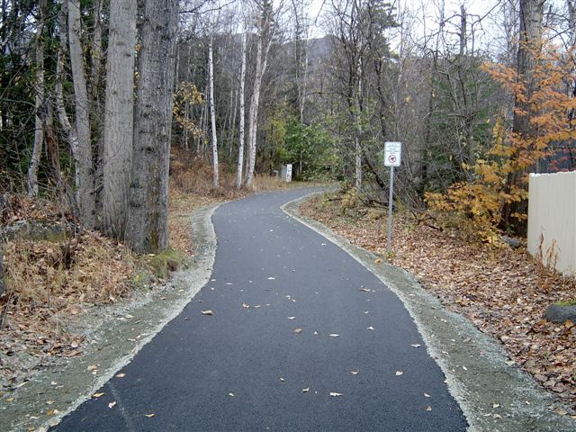 Paved pathway winding through a wooded area with trees and a sign.