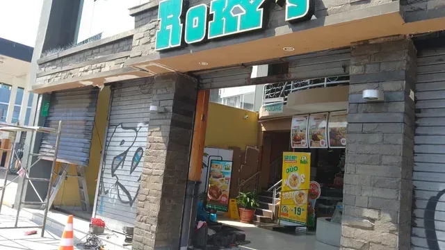 Fachada de tienda con letrero turquesa, columnas de ladrillo gris y estanterías con productos visibles en el interior.