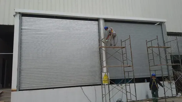 Obreros subidos a andamios instalando o reparando grandes puertas enrollables de metal gris en el exterior de un almacén.
