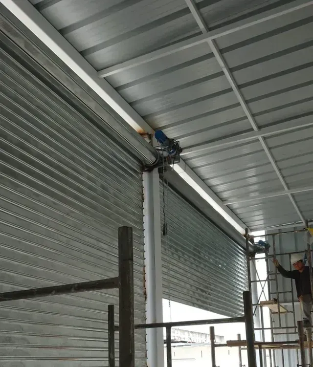 Un trabajador subido a una escalera instala una persiana enrollable de metal gris en el interior de un edificio industrial.