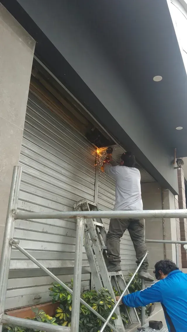 Un trabajador suelda persianas metálicas en el exterior de un edificio desde una escalera bajo una pasarela cubierta.